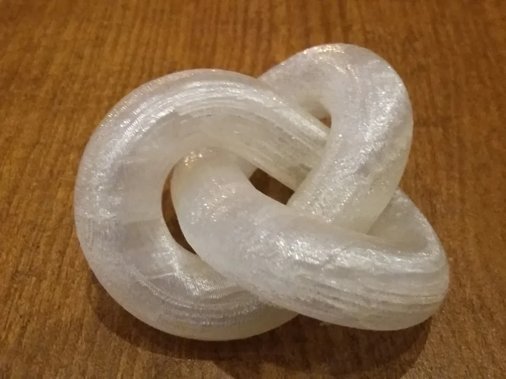 A translucent, looped 3D-printed trefoil with visible layer lines resting on a wooden surface.