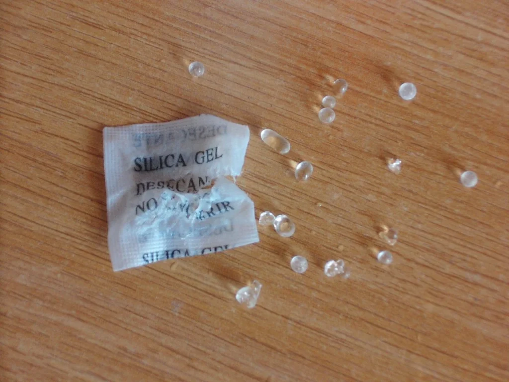 An opened silica gel packet and scattered translucent beads on a wooden surface.