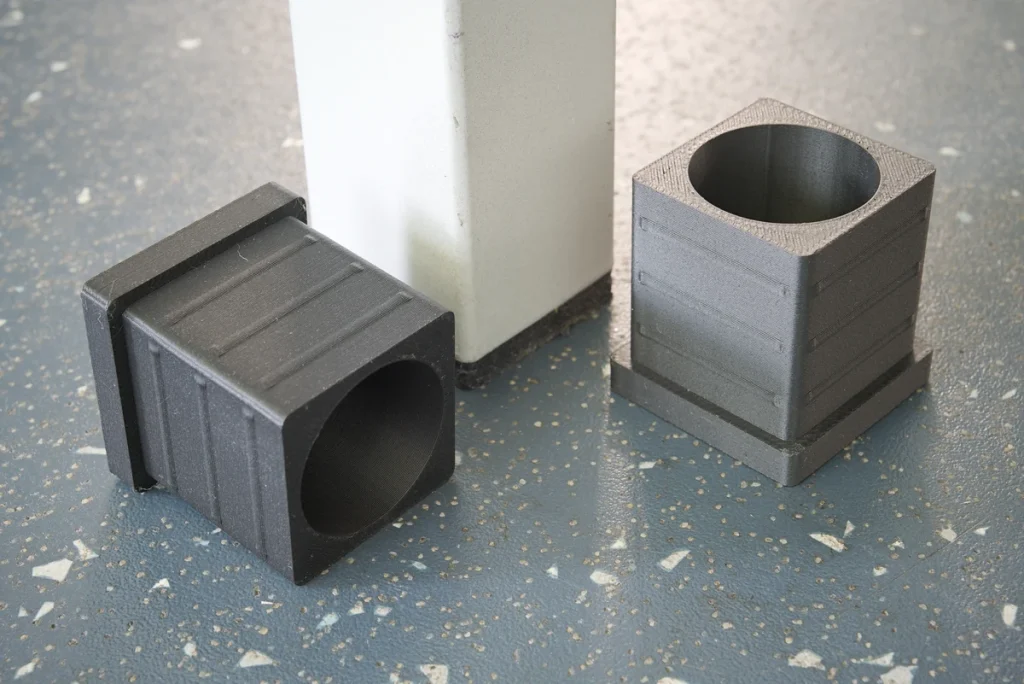 Two black 3D-printed table-leg caps with circular openings beside a white table leg on a speckled floor.
