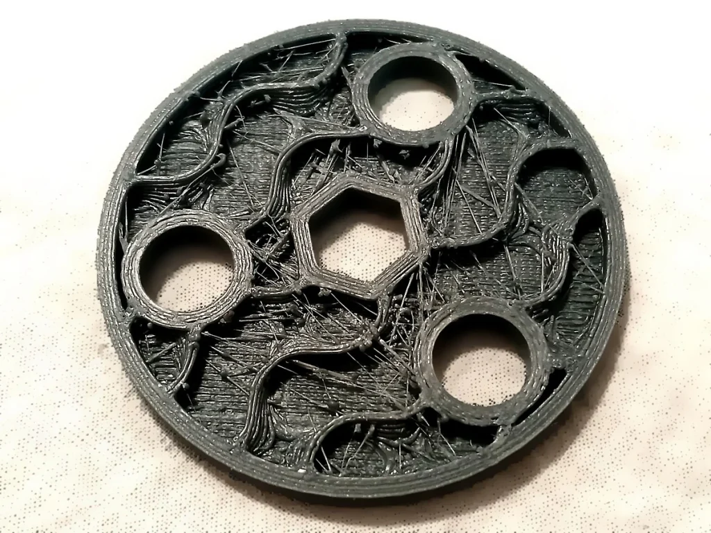 Black circular printed part with four round openings, a central hexagonal opening and thin plastic strings crossing the exposed infill.