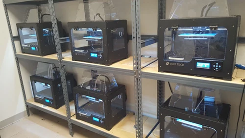Six black 3D printers with clear covers and front doors arranged on two workshop shelves.