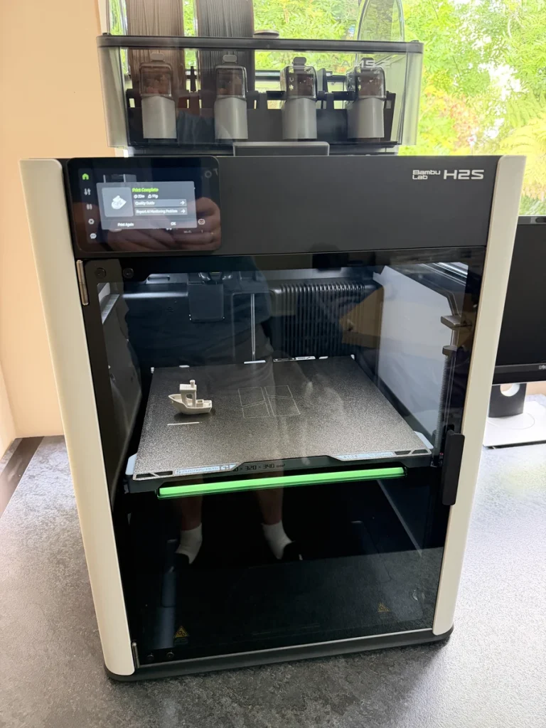Bambu Lab H2S with a closed glass front, a small printed boat on its build plate and a four-spool unit on top.