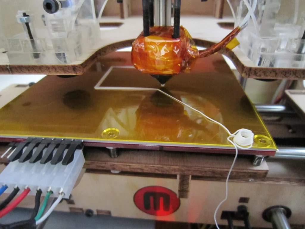 An orange-wrapped print head above a gold-colored build plate with a white square outline and loose extruded filament.