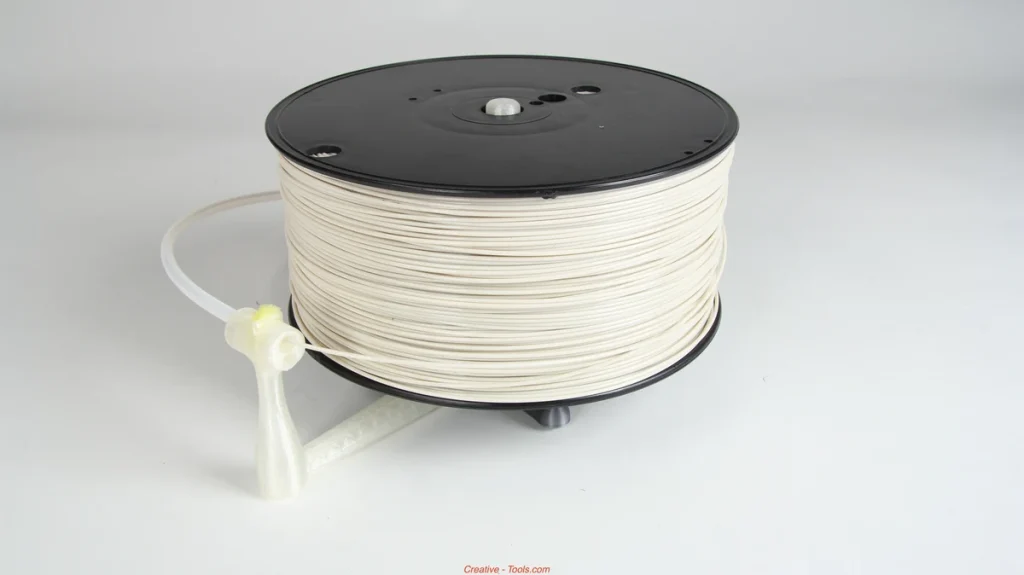 White filament on a black spool resting on a translucent printed stand, with a guide tube and small yellow pad beside it.