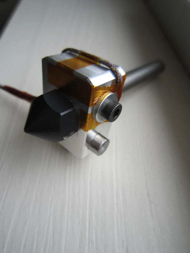 Close-up of a hot-end assembly with a dark nozzle, metal block, screws, amber tape and wires on a pale work surface.