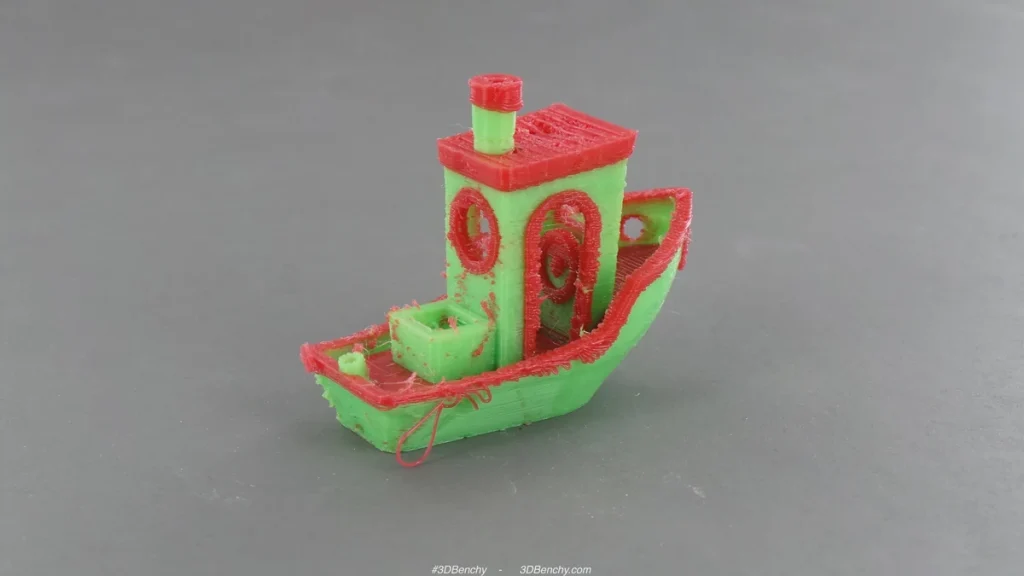 Red and green printed Benchy boat with visible strings and stray plastic on a gray surface.