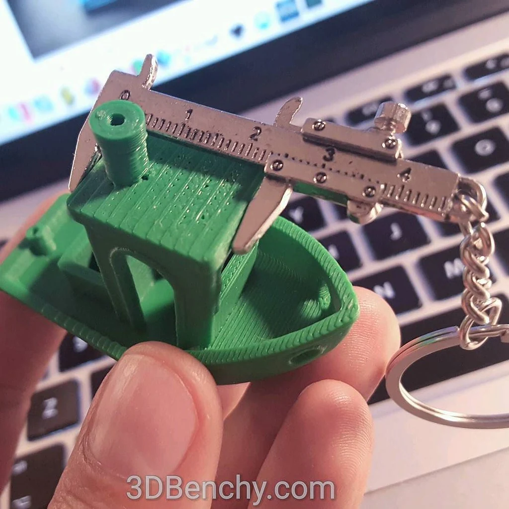 A hand holds a green printed boat while a caliper-shaped keyring spans its cabin roof, with a laptop behind it.