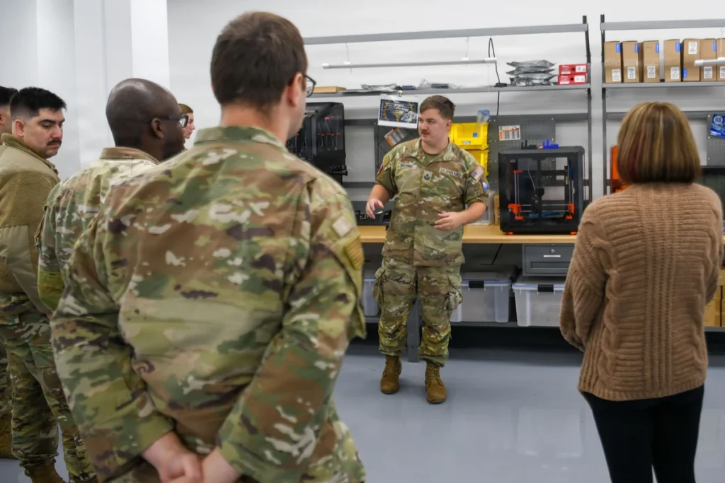 An instructor addresses workshop attendees beside black enclosed 3D printers on a workbench.
