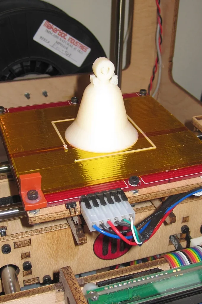 A white printed bell on a gold-taped build plate, with a MakerBot filament spool, wooden printer frame and wiring visible.
