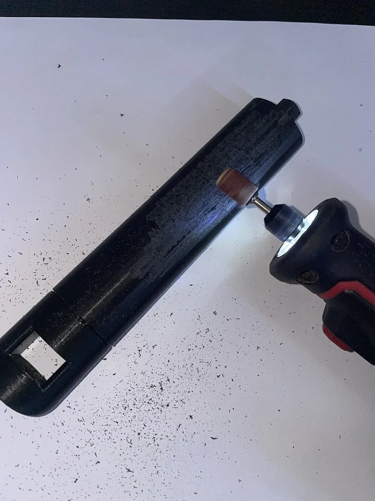 A rotary tool with a brown sanding drum touches a black printed part on a white surface scattered with dark sanding debris.