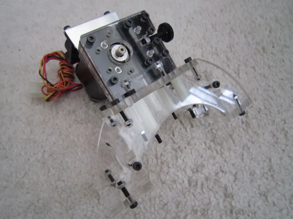Exposed filament-drive mechanism and stepper motor on a clear plastic support, resting on a textured surface.