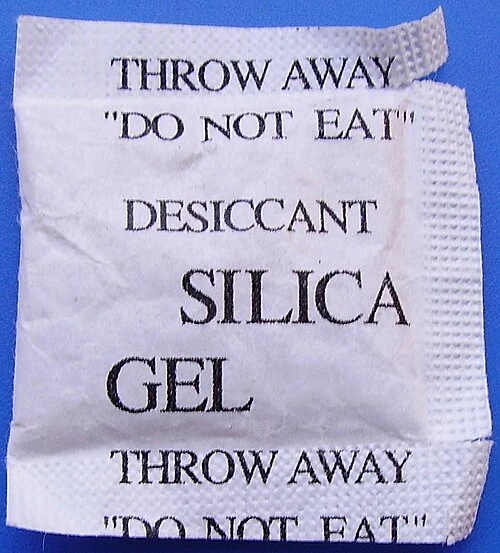 A creased white silica-gel sachet with black desiccant and do-not-eat lettering on a blue surface.