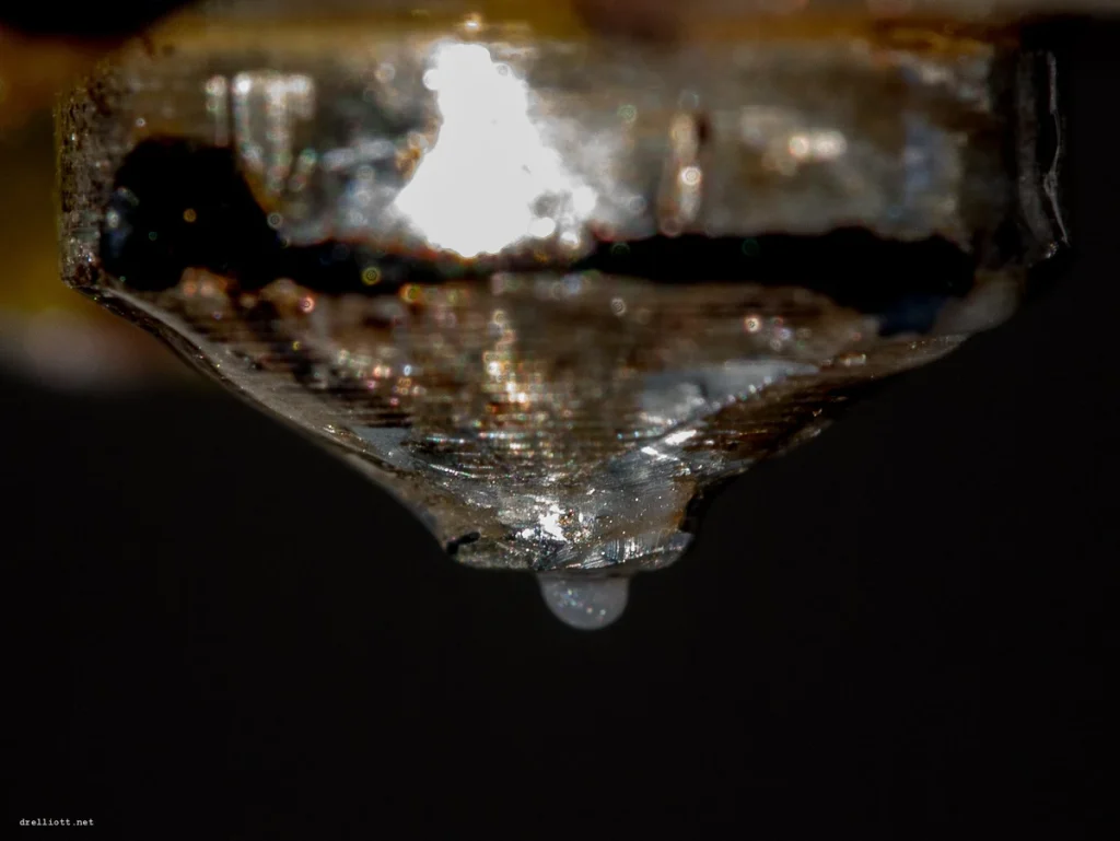 Macro photograph of a worn metal 3D printer nozzle with a small bead of plastic at its tip.