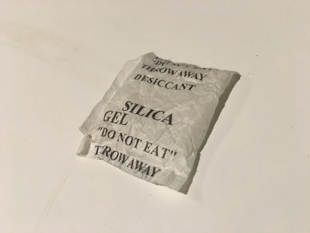 A creased white silica gel packet with black warning text rests on a pale surface and casts a shadow.