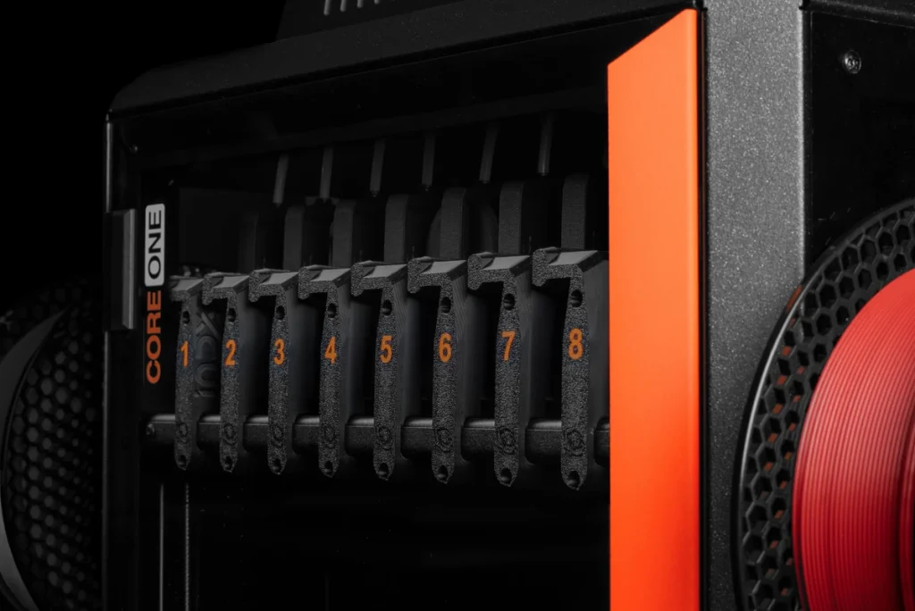 Eight numbered tool docks across the front of a CORE One INDX, beside an orange frame strip and red filament spool.