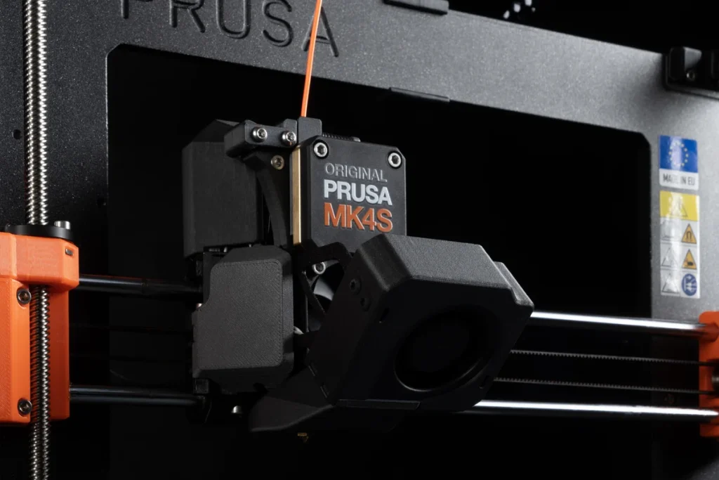Close-up of the Original Prusa MK4S printhead, forward cooling fan, orange filament and horizontal guide rods.