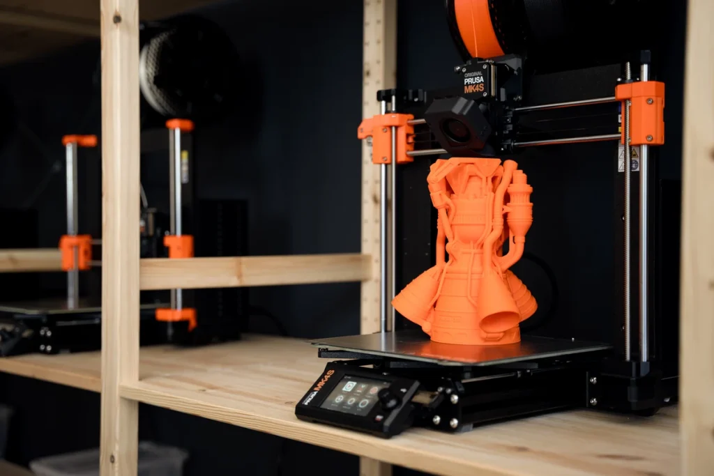 An orange rocket-engine model on the bed of a Prusa MK4S on wooden shelving.