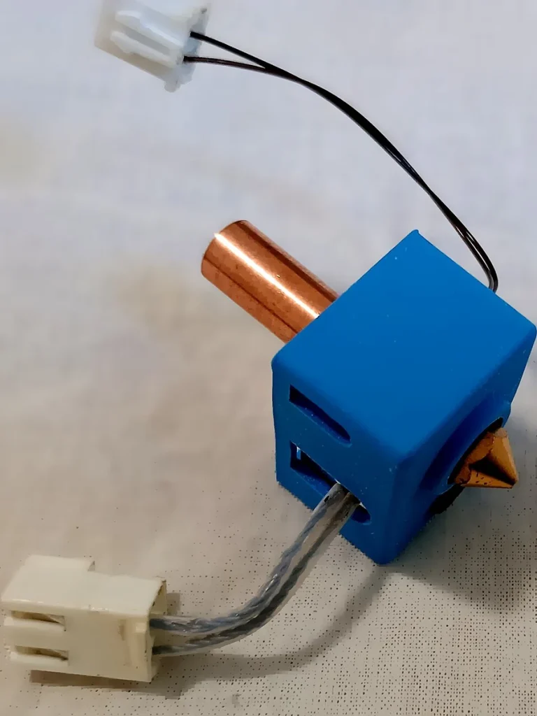 A hotend with a blue silicone cover, brass-colored nozzle, copper-colored tube and two wired white connectors lies on a pale surface.