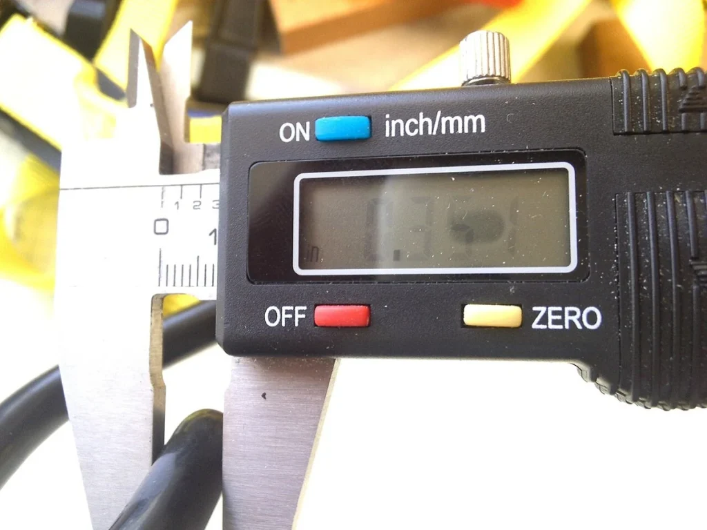 A digital caliper grips a dark curved component, with yellow straps in the background.