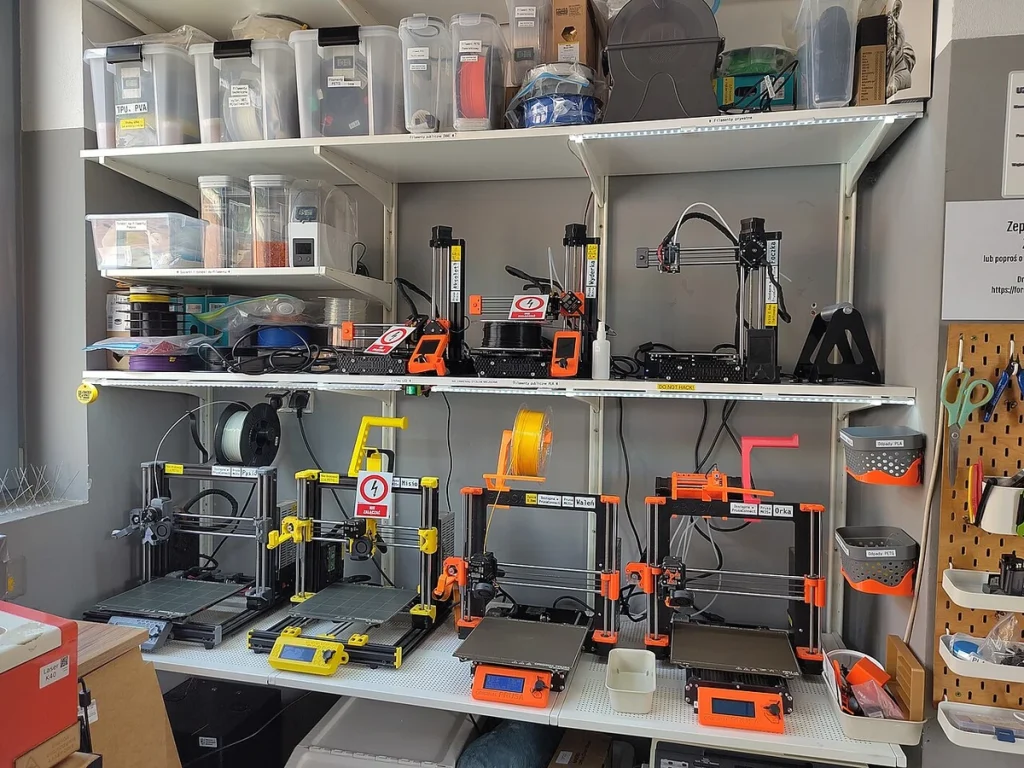Prusa printers occupy two shelves with filament containers above and hand tools hanging beside them.