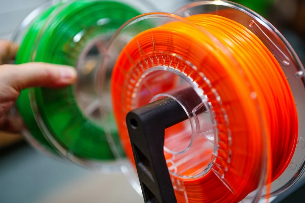 A hand touches a green filament spool behind an orange spool on a black support, both with clear plastic flanges.