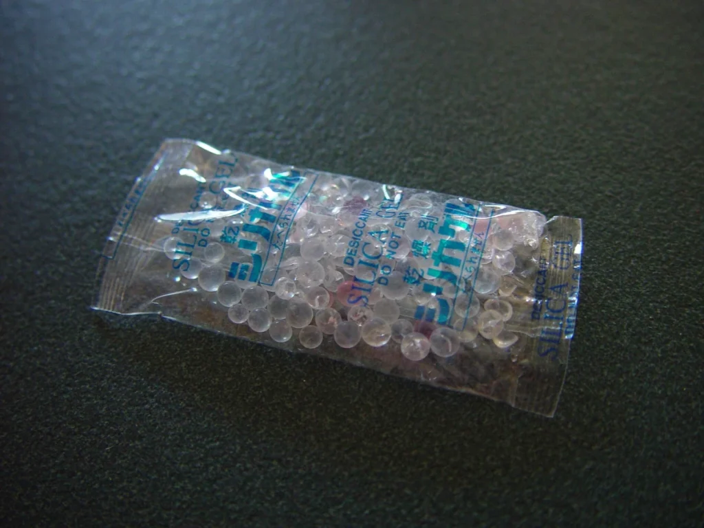 A transparent packet of silica gel beads with blue printing rests on a dark textured surface.