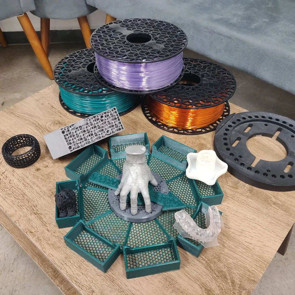 Colored filament spools sit behind perforated trays, a hand-shaped model and other printed objects on a wooden table.