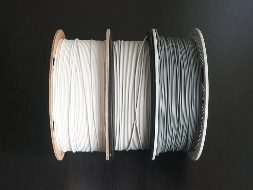 Three filament rolls in white, light gray and dark gray stand side by side on a dark surface.
