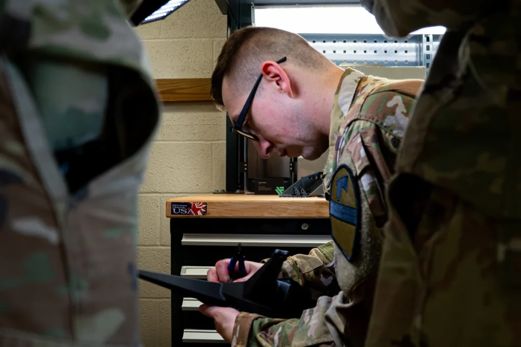 A uniformed technician wearing glasses uses blue-handled cutters on a black printed aircraft frame at a workbench.